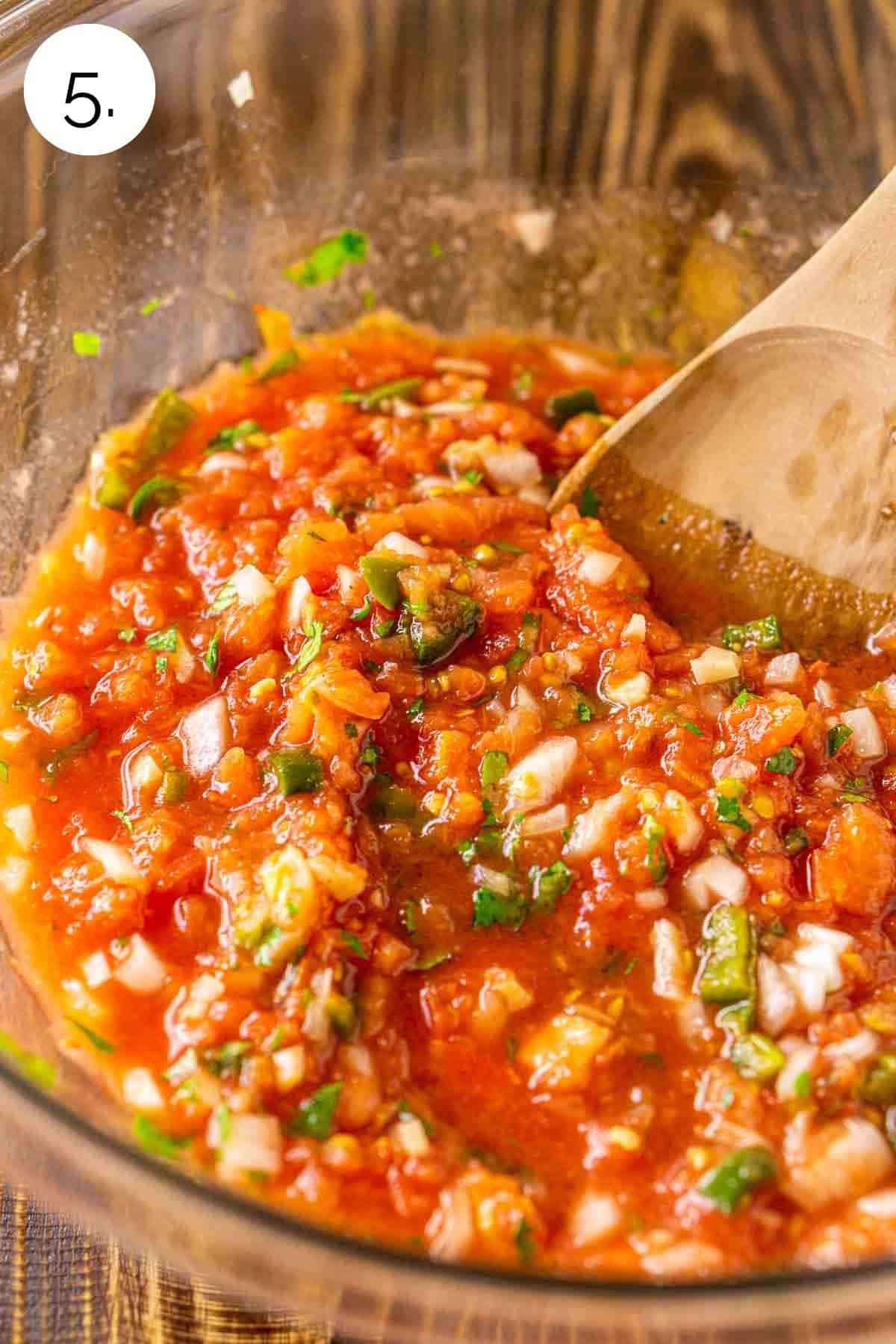 A brown wooden spoon stirring together the smoked tomatoes with the remaining ingredients in a large glass bowl.