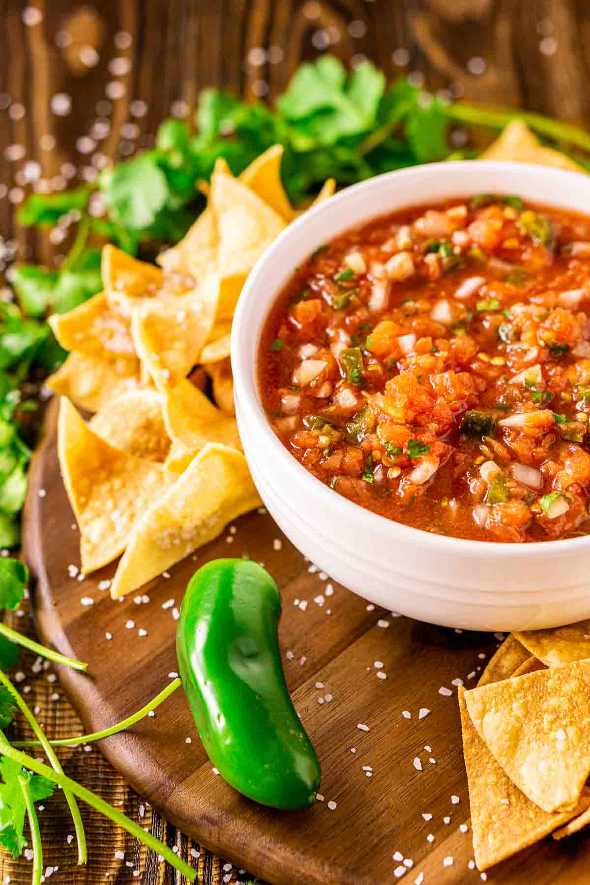 A white bowl of the smoked salsa on a brown serving tray with chips and salt all around it and cilantro sprigs and a jalapeño to the side.