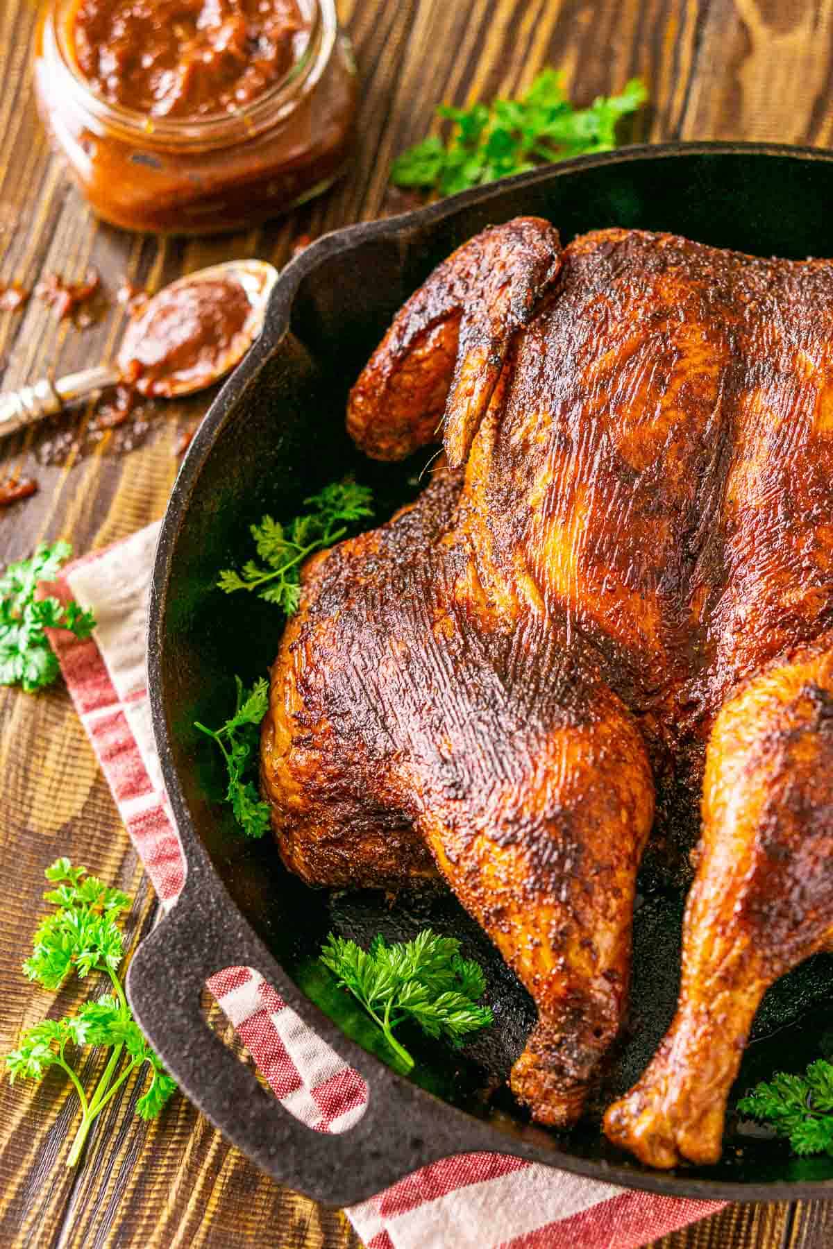 A side view of the smoked spatchcock chicken in a black cast-iron skillet on top of a red and white napkin with parsley sprinkled around it.