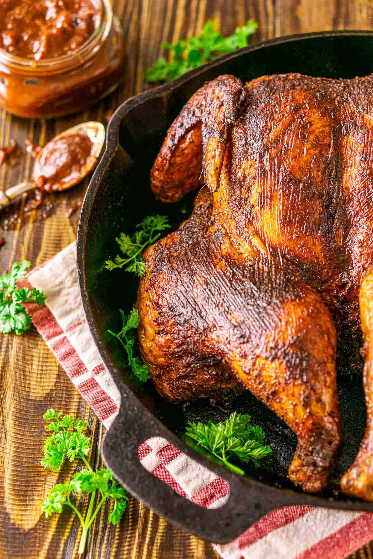 The smoked spatchcock chicken in a black cast-iron skillet on a brown wooden surface with sprigs of parsley to the left.