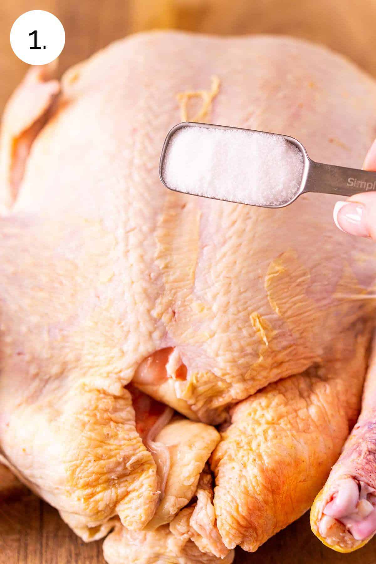 A hand holding a tablespoon over the chicken on a brown cutting board before dry brining the skin.