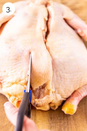 The chicken on a brown wooden cutting board with the kitchen shears making the second cut along the backbone on the left side.