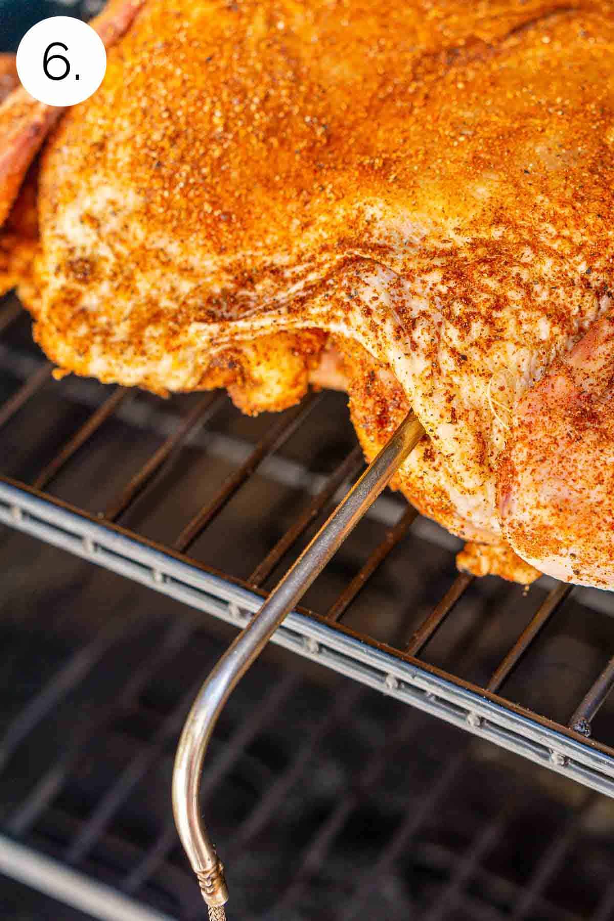 The chicken on the smoker grill grates with a leave-in meat thermometer inserted into the breast meat.