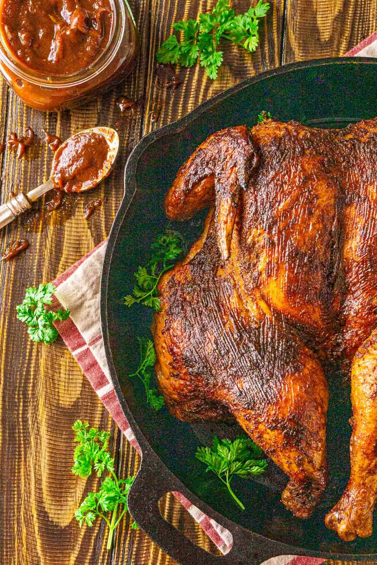 An aerial view of the smoked spatchcock chicken in a black cast-iron skillet with a jar of BBQ sauce to the left.