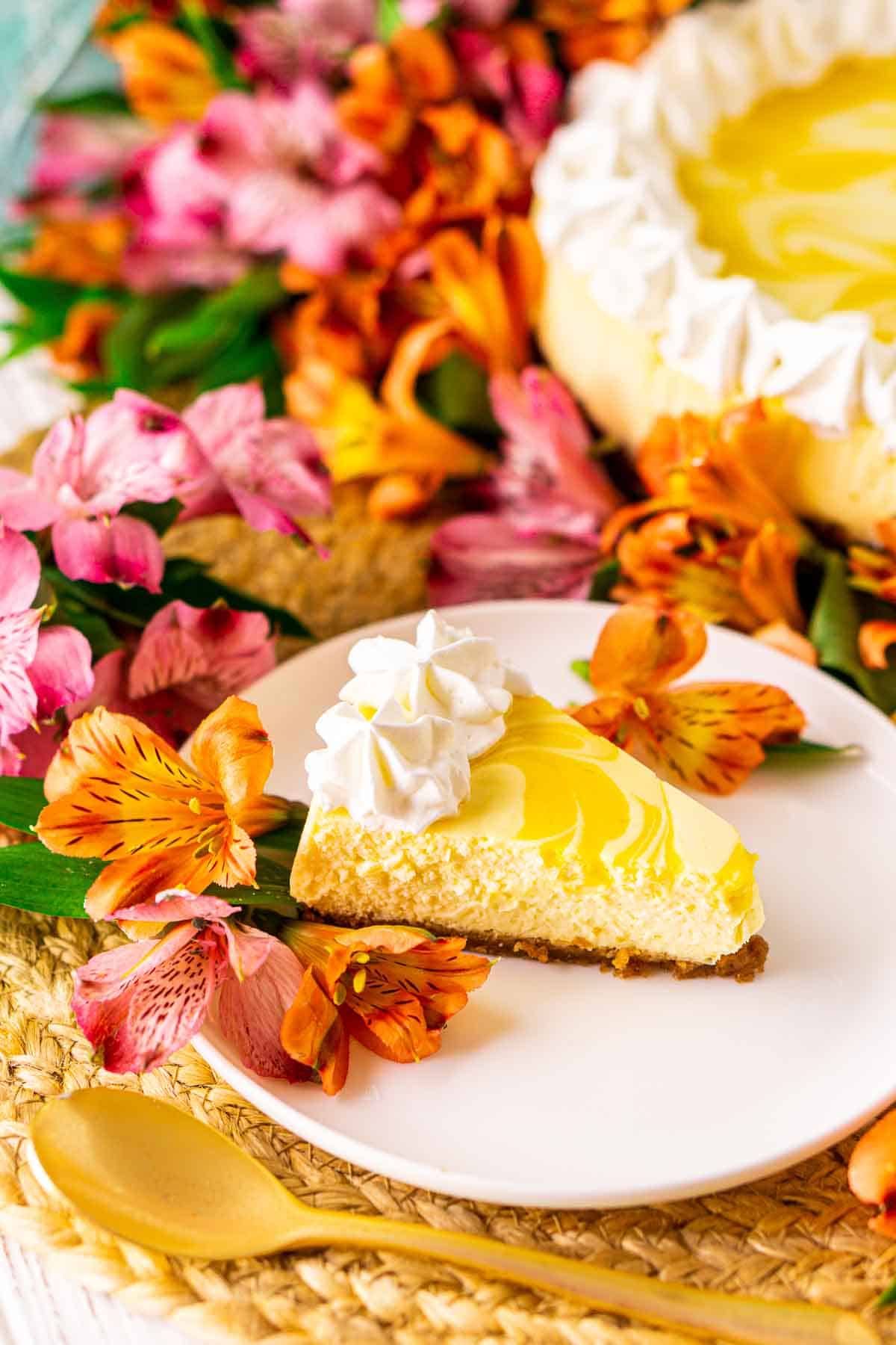 A slice of passion fruit cheesecake on a white plate with a gold spoon in front and tropical flowers around it.