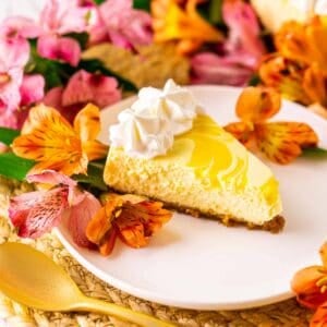 Looking straight on to a slice of passion fruit cheesecake on a white plate and straw placemat with colorful flowers behind it.