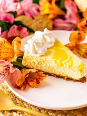 Looking straight on to a slice of passion fruit cheesecake on a white plate and straw placemat with colorful flowers behind it.