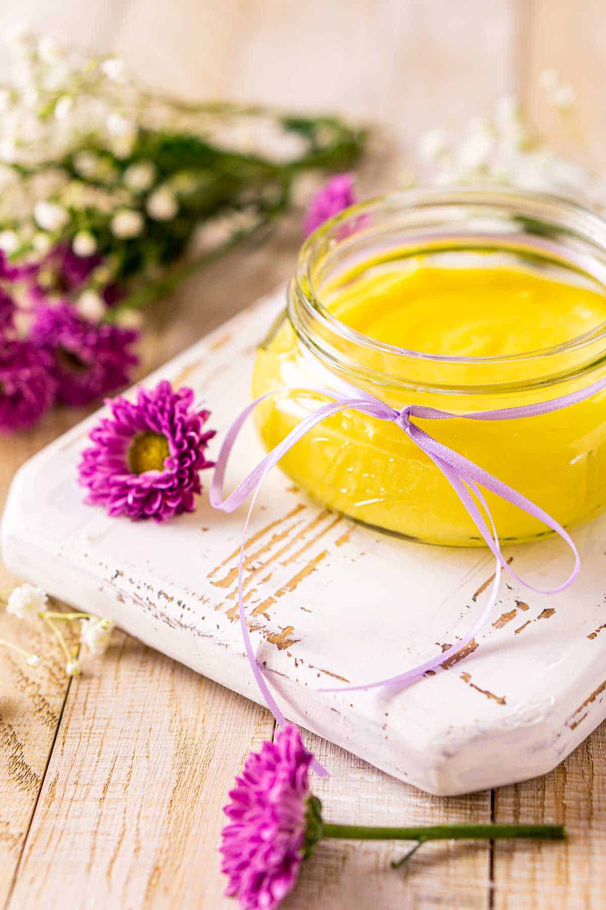 Looking straight on to a glass jar of passion fruit curd on a white serving tray with a purple flower in front.