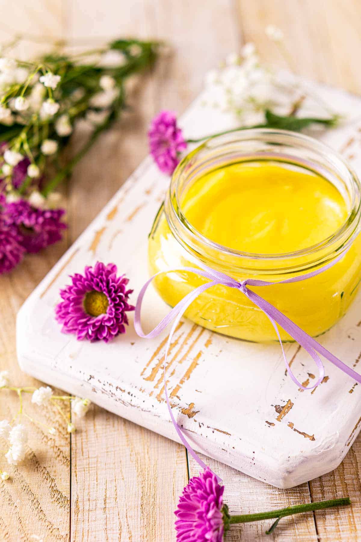 A glass jar of passion fruit curd on a white serving tray with purple and white flowers on a cream-colored wooden surface.