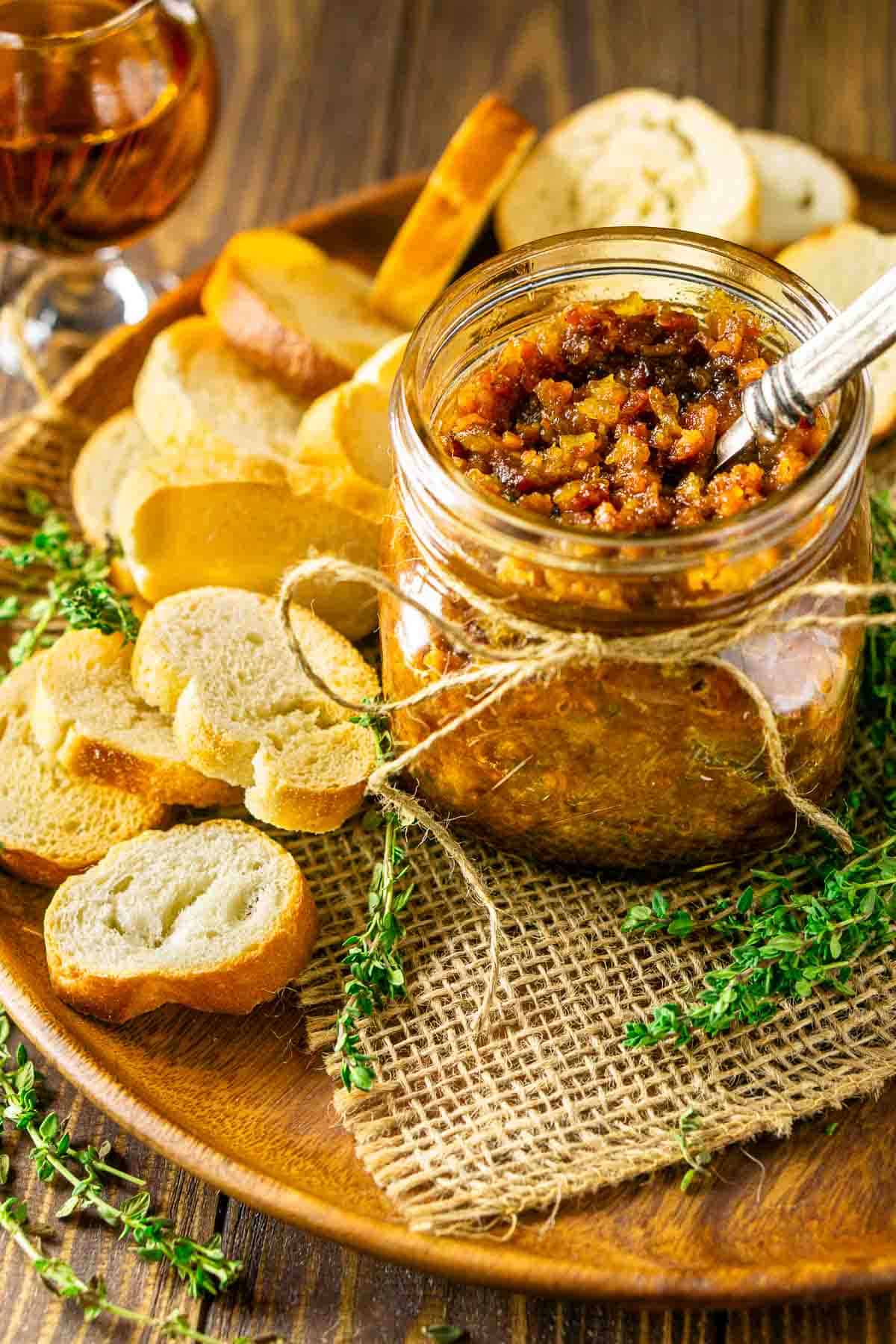 A small mason jar on burlap filled with bourbon bacon jam and a pile of bread rounds and glass of bourbon next to it.