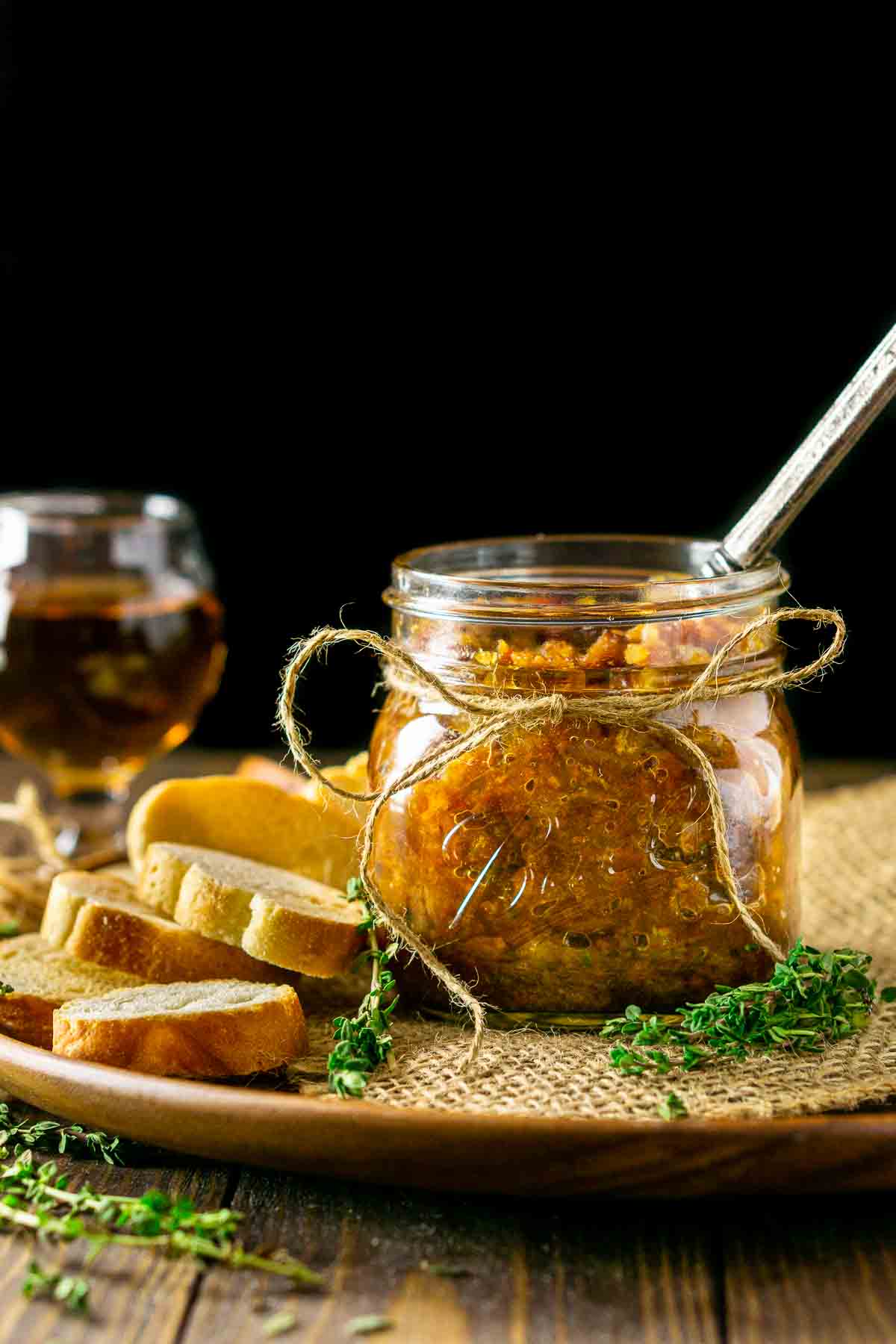 A jar of bourbon bacon jam with a spoon sticking out of it and a small glass of bourbon behind it against a black background.