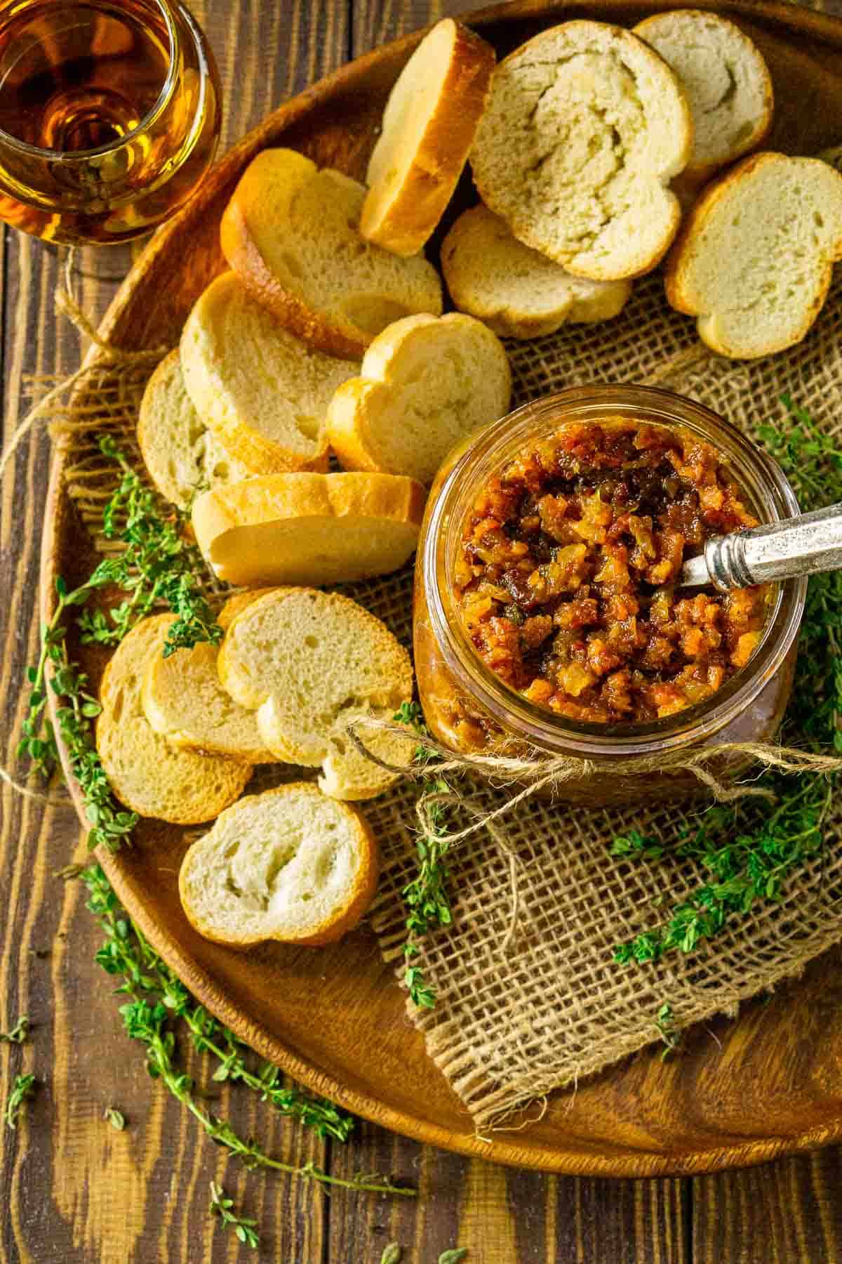 An aerial view of a jar of bourbon bacon jam on a wooden brown plate with a glass of bourbon in the upper lefthand corner.