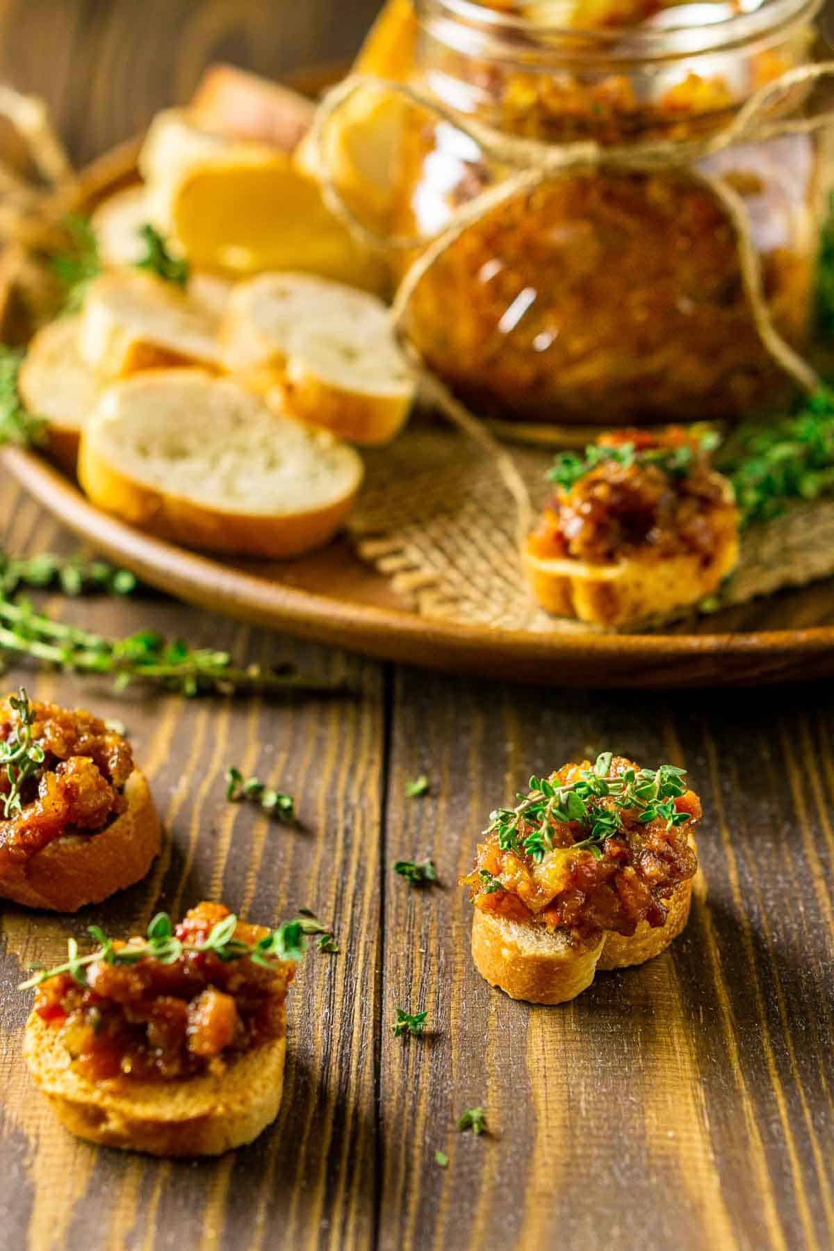 The bourbon bacon jam spooned onto the top of crostini with a jar of the jam on a wooden plate in the background.