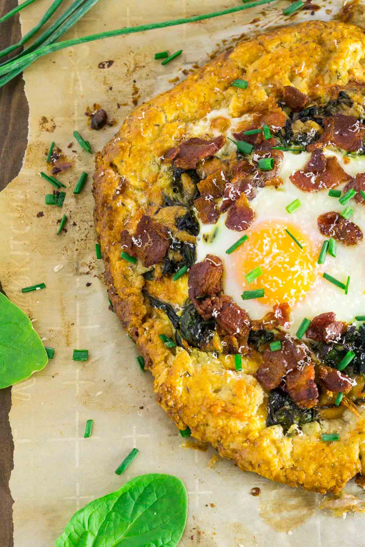 The breakfast galette on brown parchment paper before it's been cut with pieces of spinach to the side.