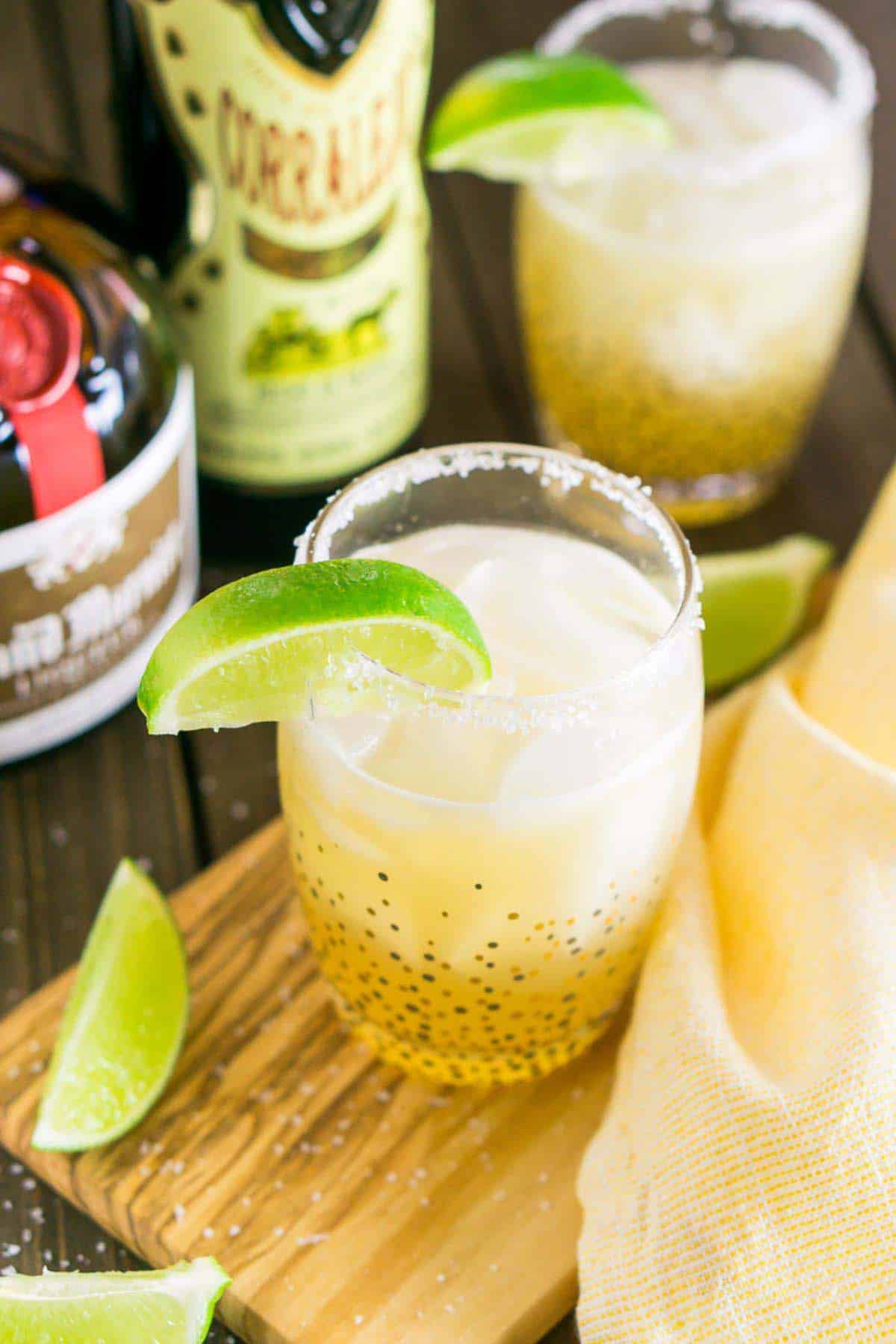 A classic margarita on a small wooden serving tray with a yellow napkin to the side and lime slices to the right.