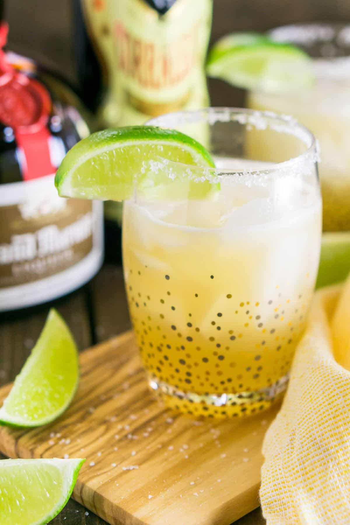 A close-up shot of a classic margarita on a wooden serving tray with salt scattered around and lime slices to the side.