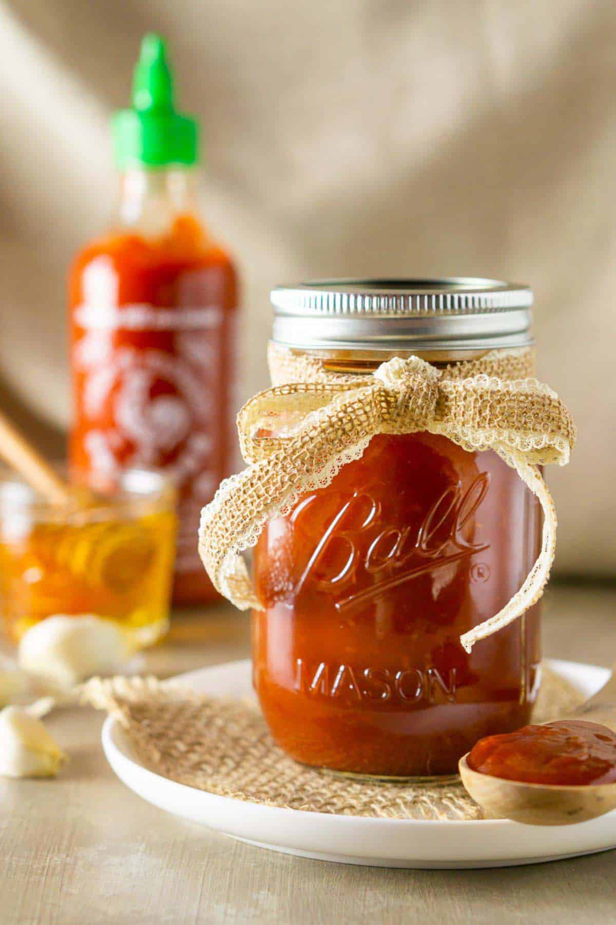 A glass mason jar of the honey-sriracha BBQ sauce on a white plate with burlap and a wooden spoon full of the sauce on the right.