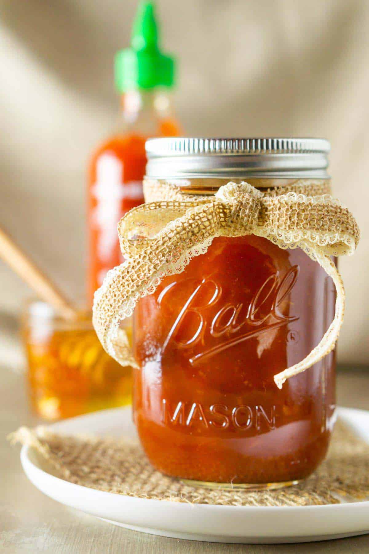 A straight-on shot of a glass jar filled with the honey-sriracha BBQ sauce on a piece of burlap on top of a small white plate with a jar of honey in the background.