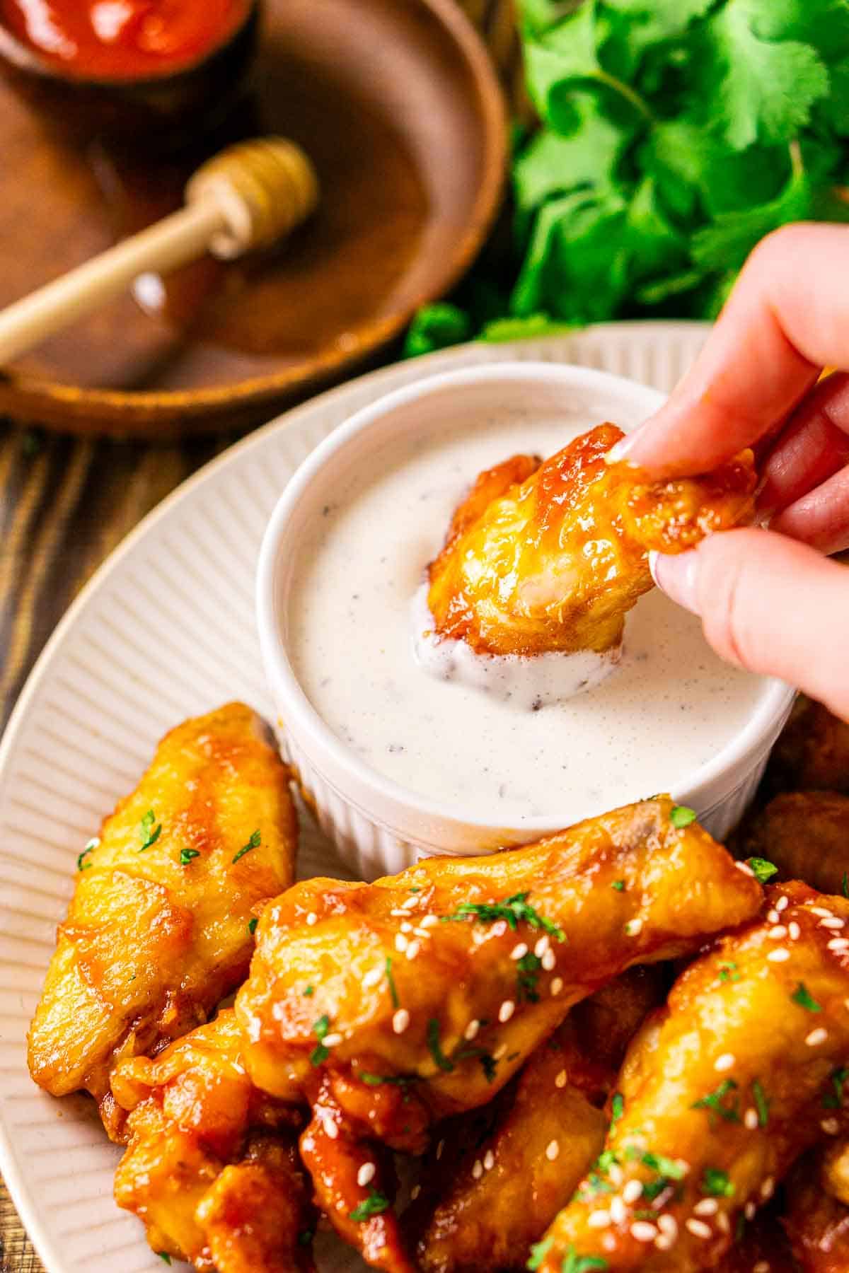A hand dipping a honey-sriracha wing in a white bowl filled with ranch dressing and a bundle of fresh cilantro in the background.