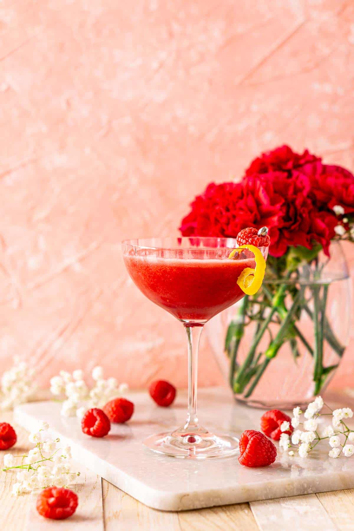 Looking straight on to the raspberry martini on a white marble tray with white flowers and fresh raspberries around it.