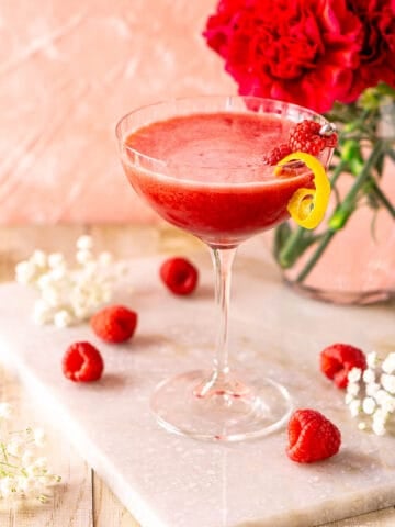 A fresh raspberry martini on a white marble tray with a bouquet of hot pink flowers behind it and white flowers and raspberries placed around it.
