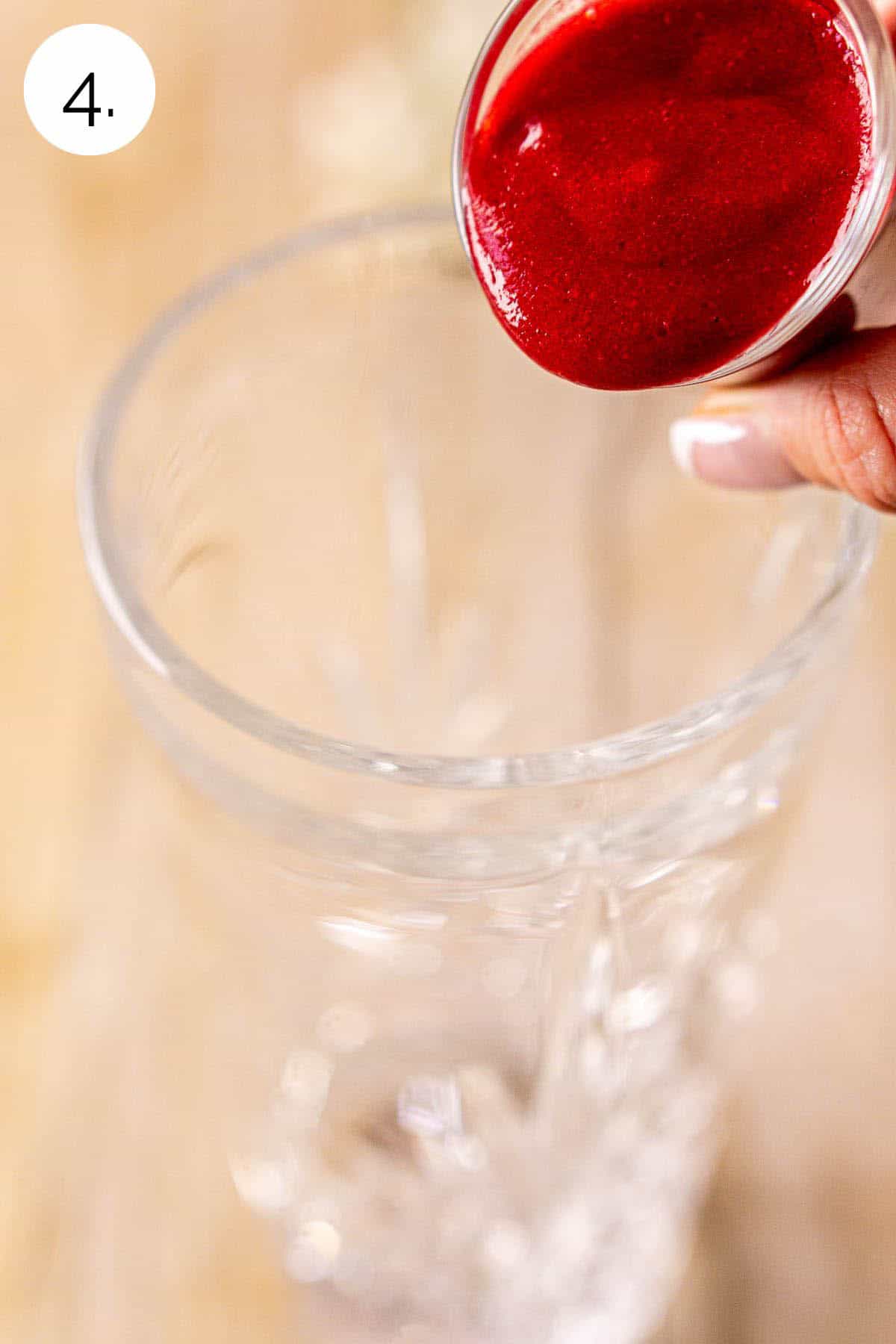 A hand pouring a shot glass of the raspberry purée into a crystal cocktail shaker against a cream-colored wooden surface.