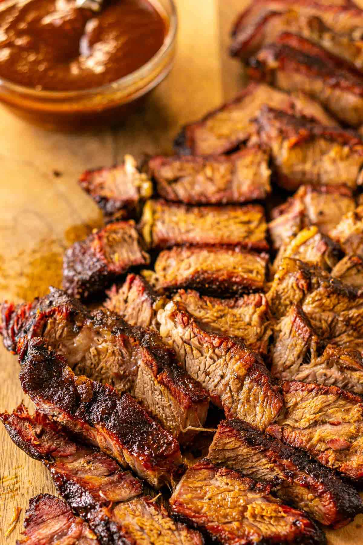 A close-up view of the smoked chuck roast cut into slices on a brown cutting board with a bowl of BBQ sauce in the upper lefthand corner.