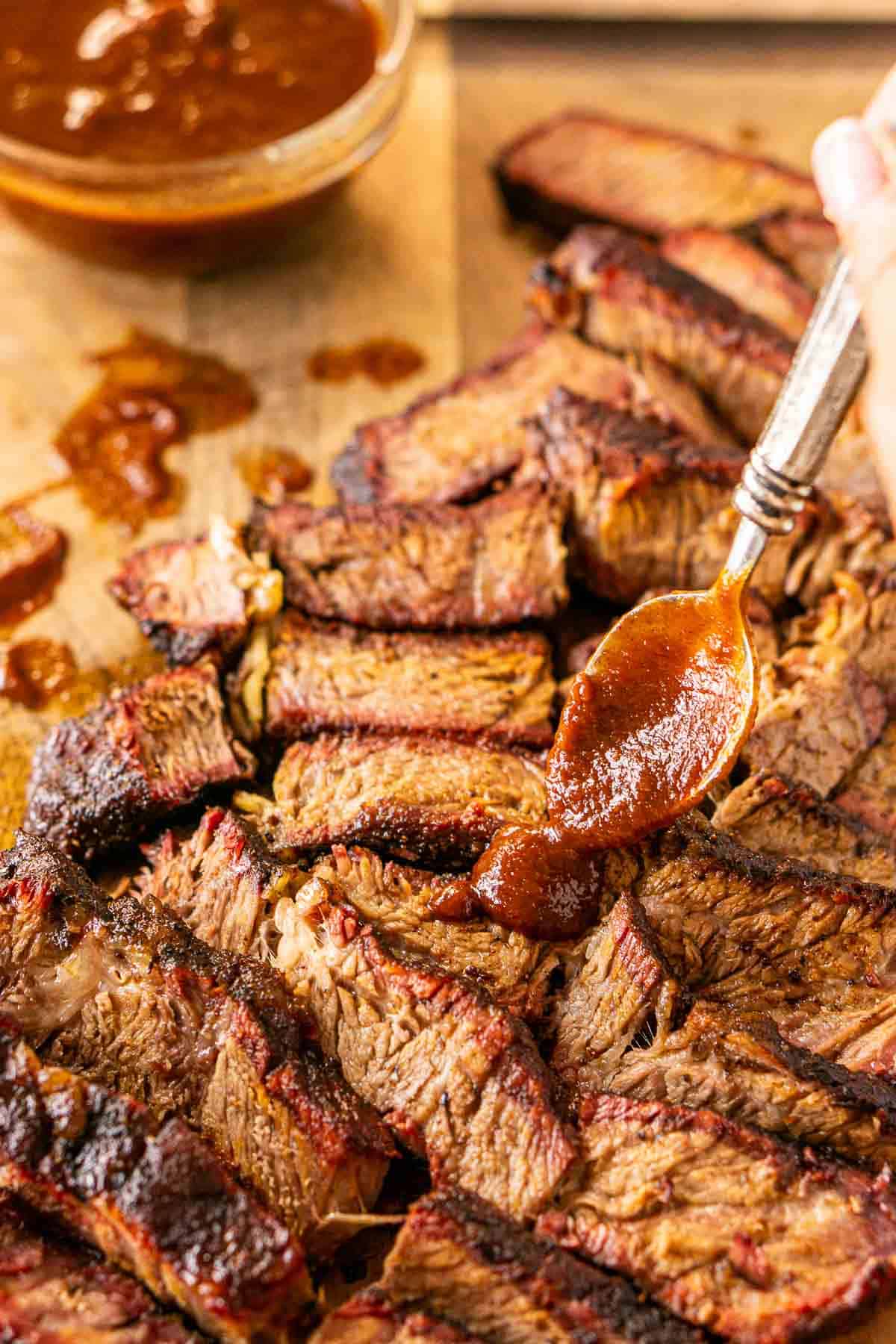 A silver spoon adding some BBQ sauce on top of the slices of smoked chuck roast on a brown wooden cutting board.