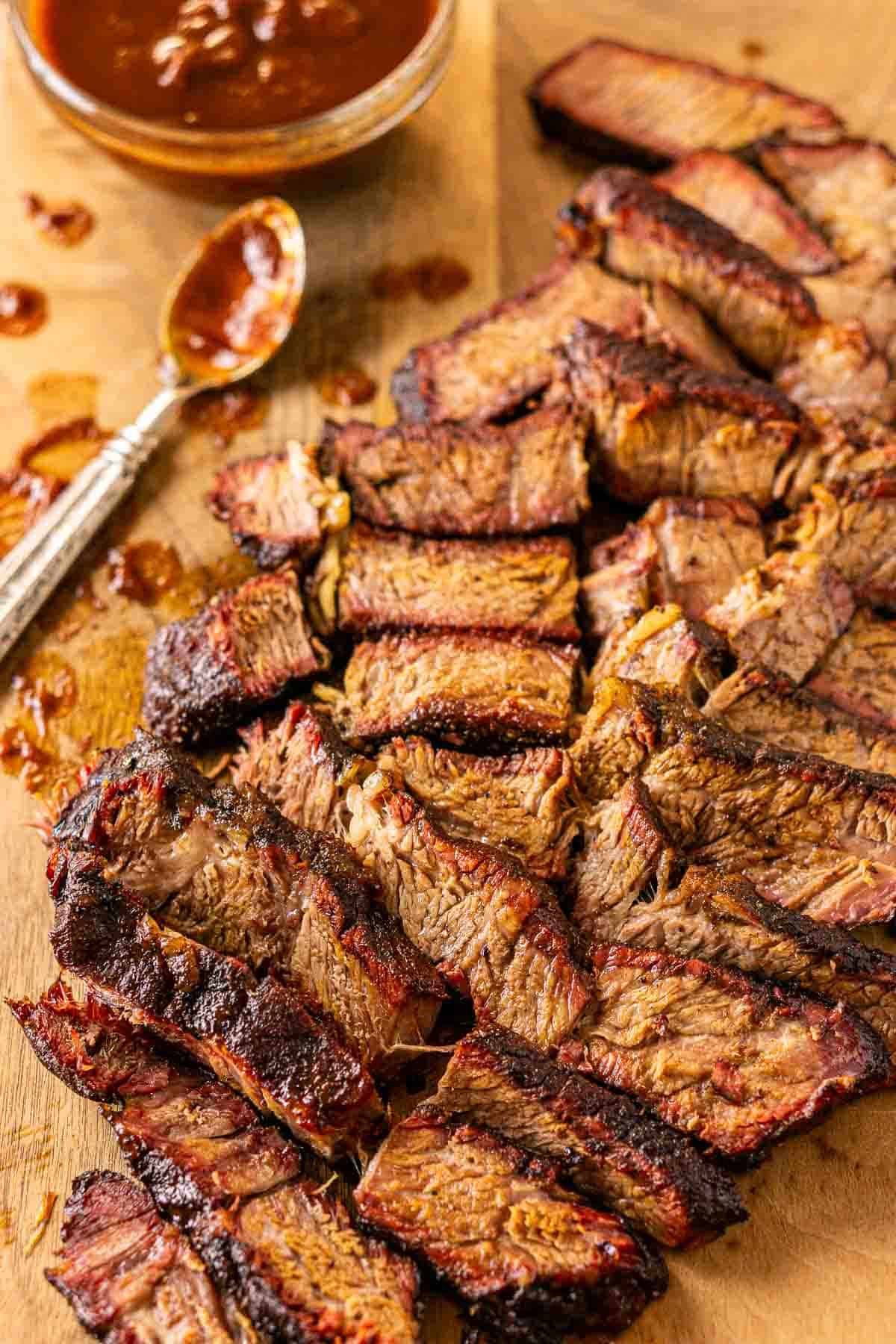 The sliced smoked chuck roast on a brown cutting board with a spoon of BBQ sauce in the top-left corner.