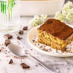 A close-up of a slice of chocolate tiramisu on a white plate with a silver spoon in front and chopped chocolate around them.