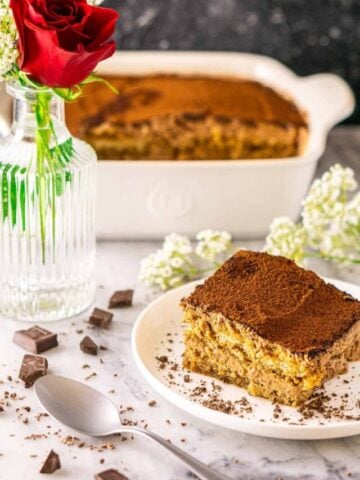 A slice of chocolate tiramisu on a small white plate with a rose in a vase to the left and chopped chocolate scattered around it.