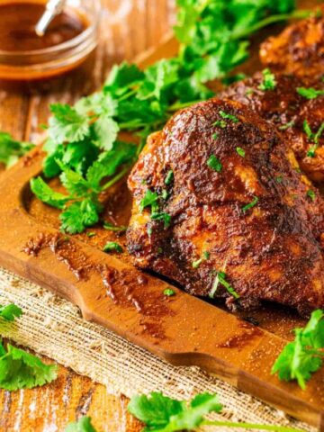 The smoked chicken breast on a brown wooden tray surrounded by sprigs of parley and a bowl of BBQ sauce in the background.
