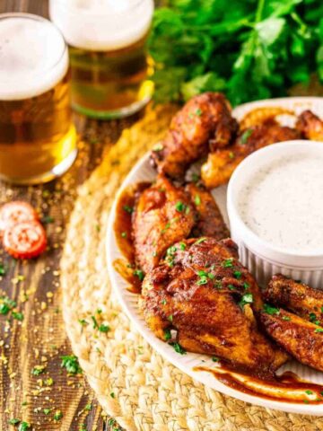 A white plate with smoked chicken wings and a bowl of ranch dressing on a straw placemat with two beers in the background and chopped parsley to the left.