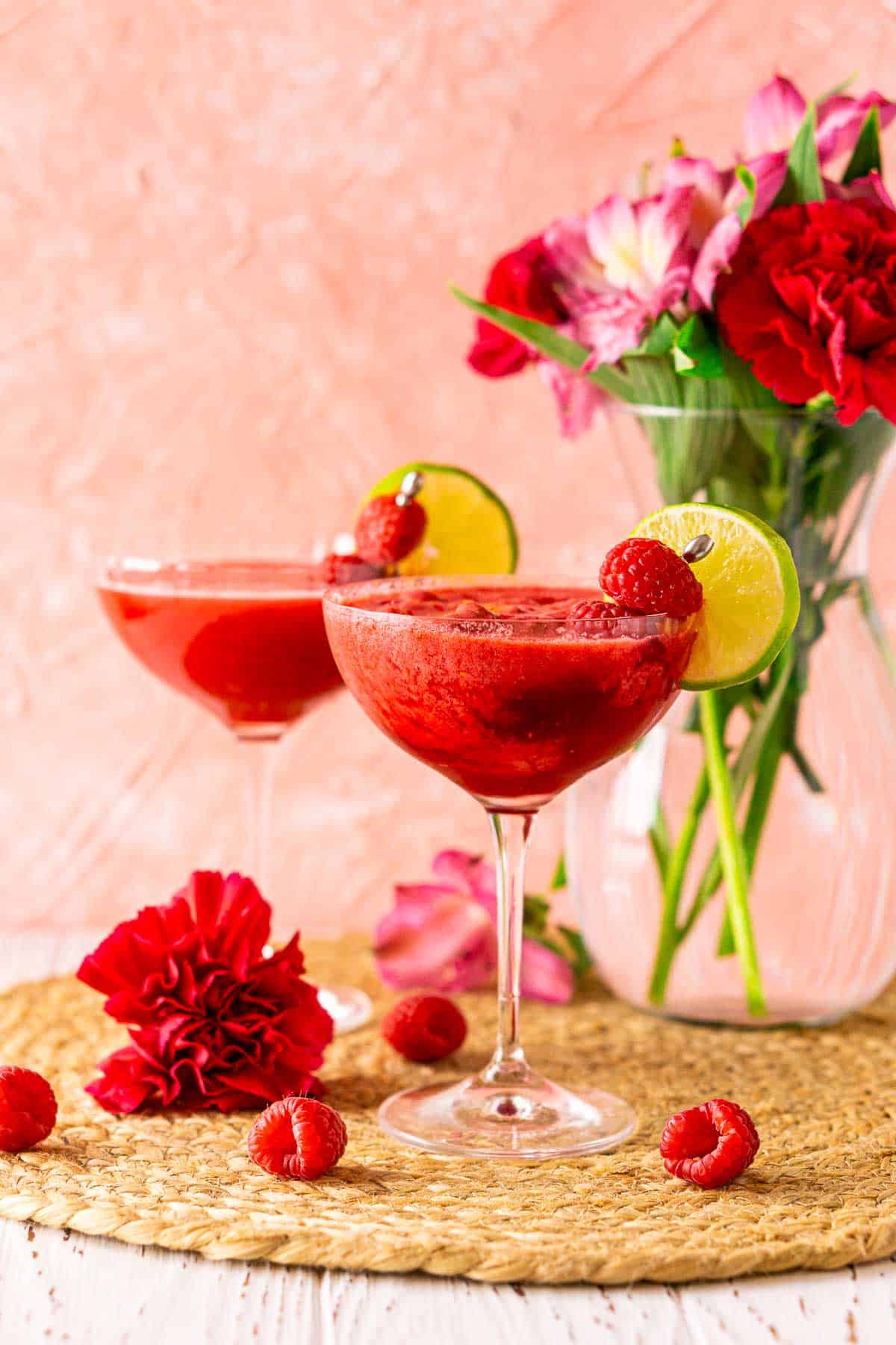 Looking straight on to two shaken raspberry daiquiris on a straw placemat over a white wooden surface with flowers around them.
