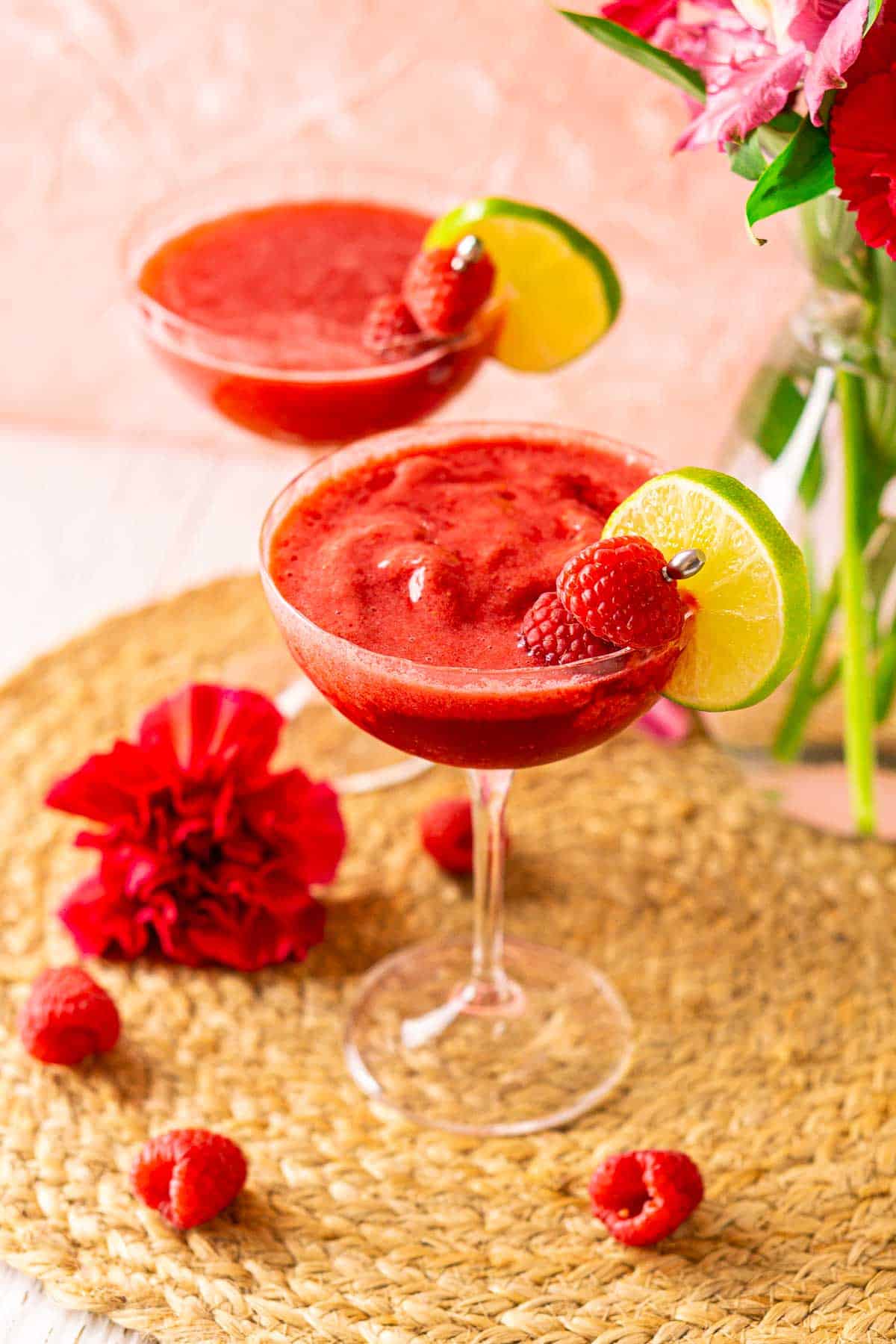 Looking down on two raspberry daiquiris on a straw placemat with fresh raspberries and pink flowers around them.