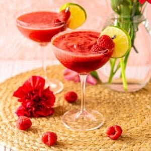 A frozen raspberry daiquiri on a straw placemat against a pink background with flowers and fresh raspberries surrounding it.