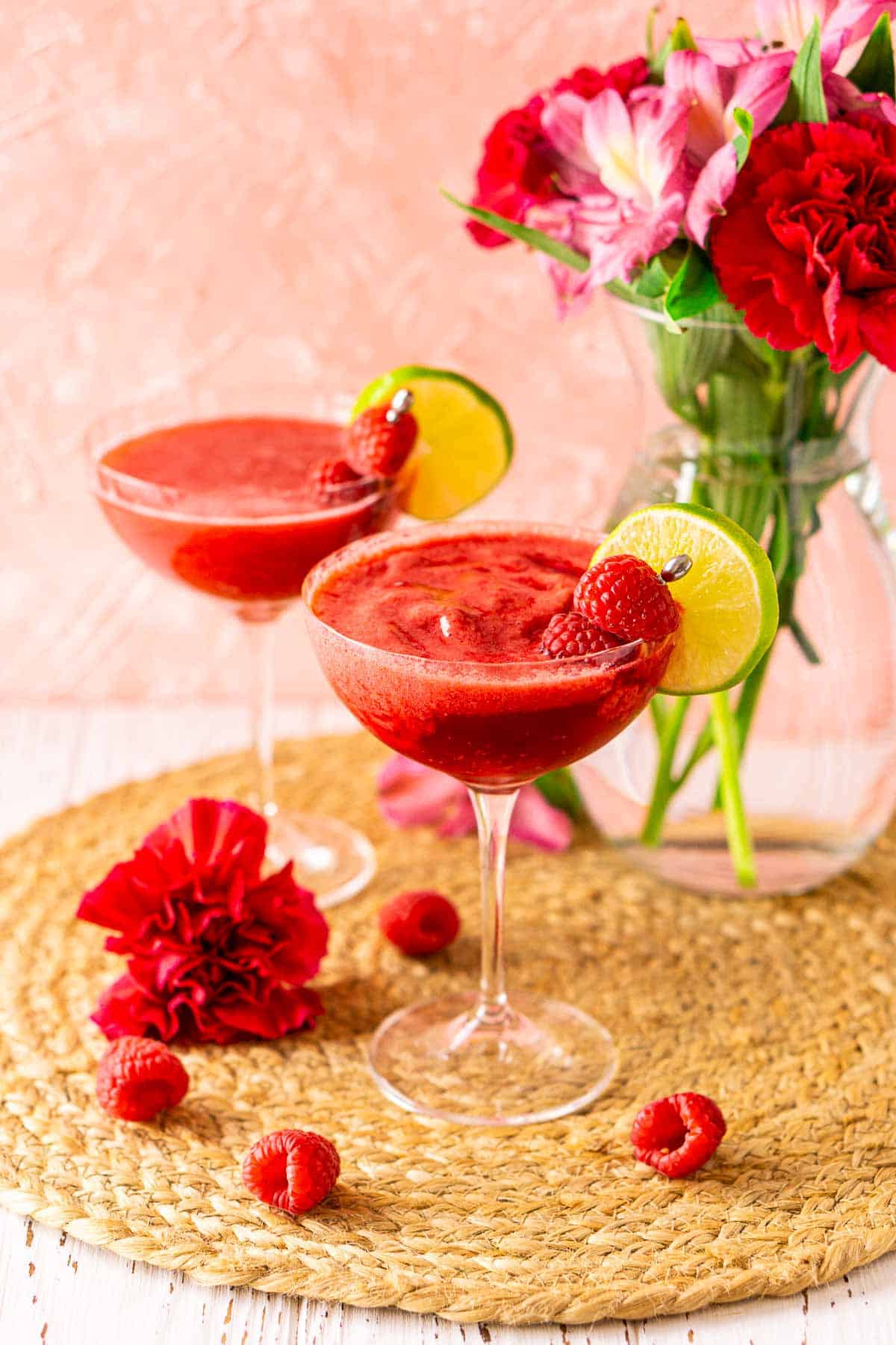 A frozen raspberry daiquiri on a straw placemat against a pink background with flowers and fresh raspberries surrounding it.
