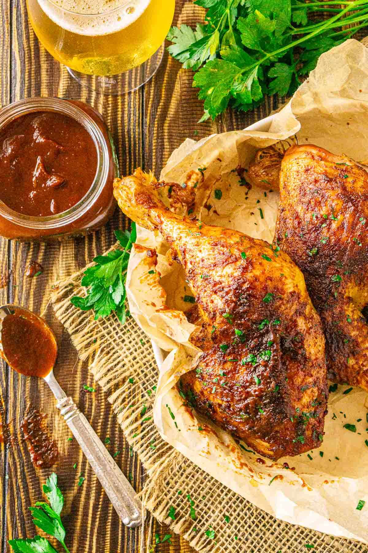 An aerial shot of the smoked chicken leg quarters on a parchment paper-lined white dish with BBQ sauce and a beer to the left.