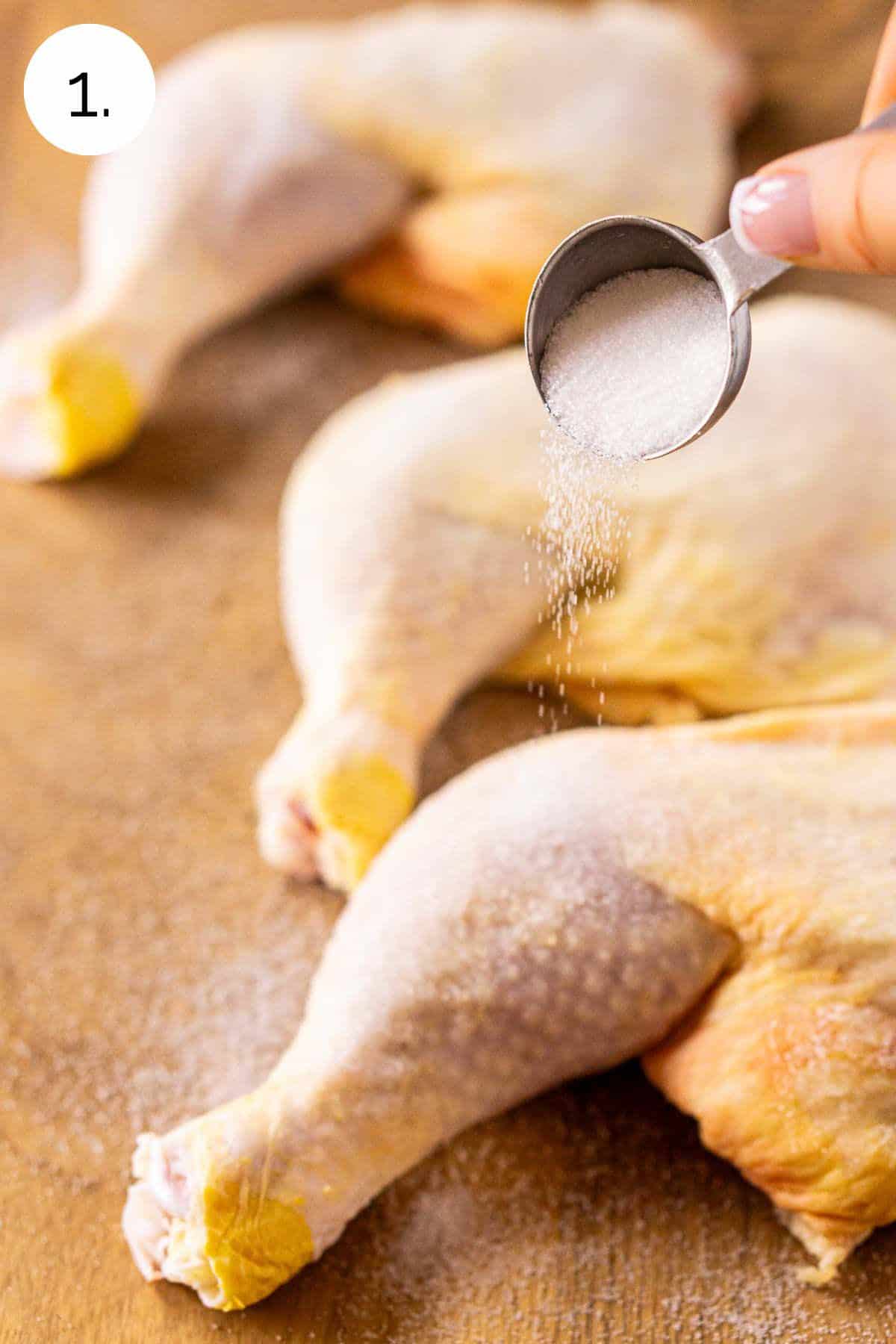 A hand sprinkling salt from a tablespoon onto the chicken quarters on top of a brown wooden cutting board.