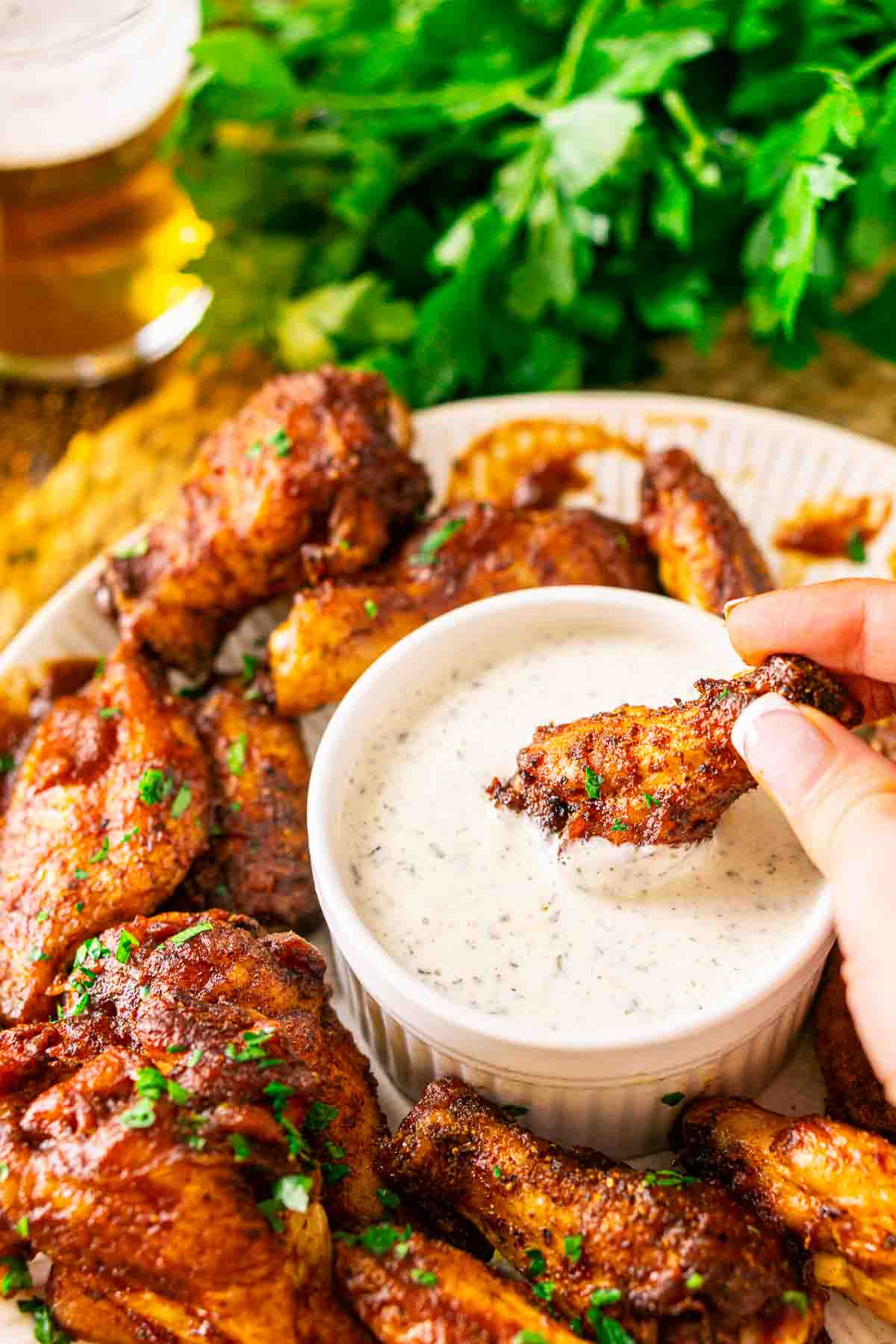 A hand dipping a smoked chicken wing into a small white bowl filled with ranch dressing with a beer in the background.