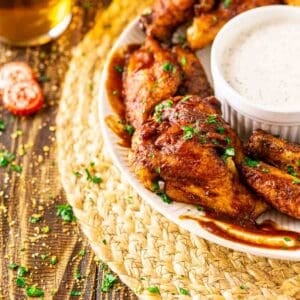 A white plate with smoked chicken wings and a bowl of ranch dressing on a straw placemat with chopped parsley to the left.