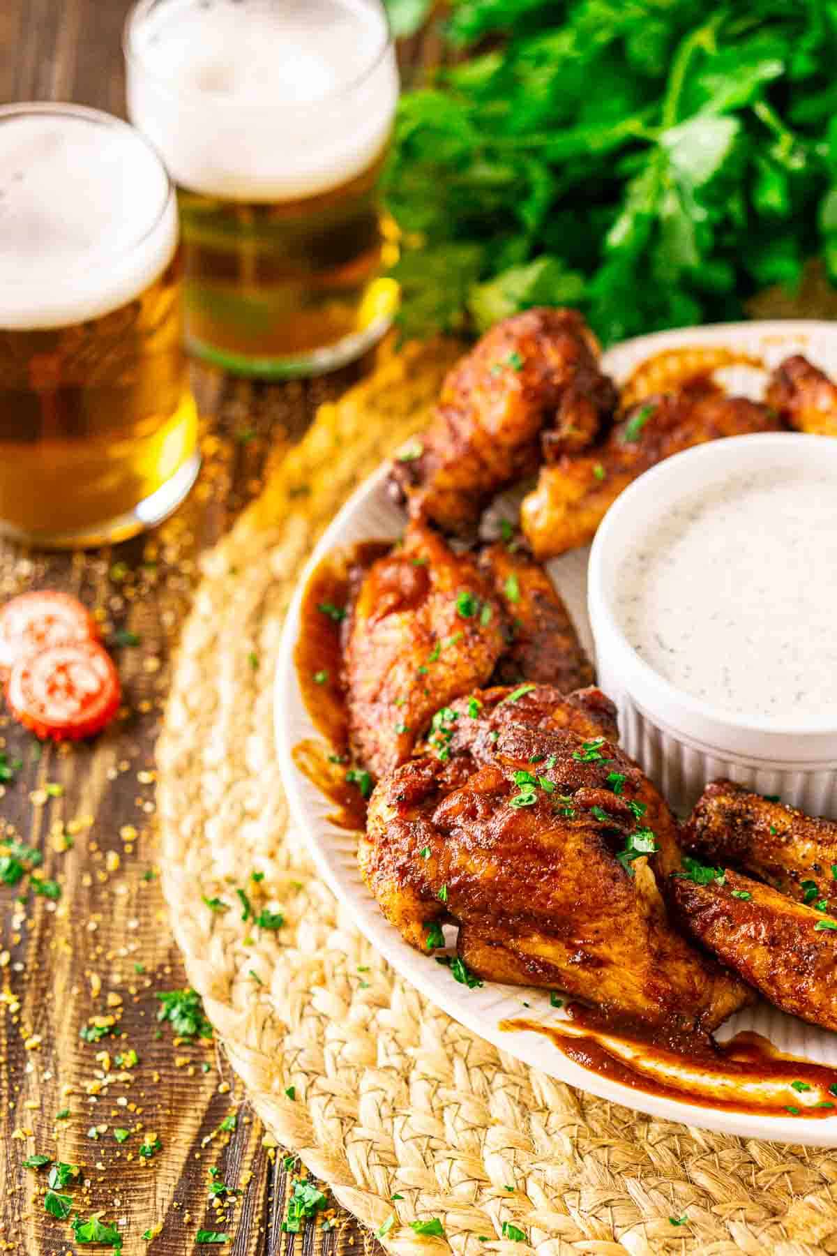 A white plate with smoked chicken wings and a bowl of ranch dressing on a straw placemat with two beers in the background and chopped parsley to the left.