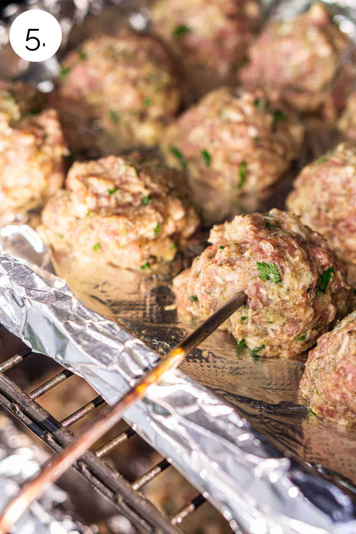 The meatballs on a baking sheet lined with aluminum foil in the smoker with a leave-in meat thermometer inserted into one of the meatballs.