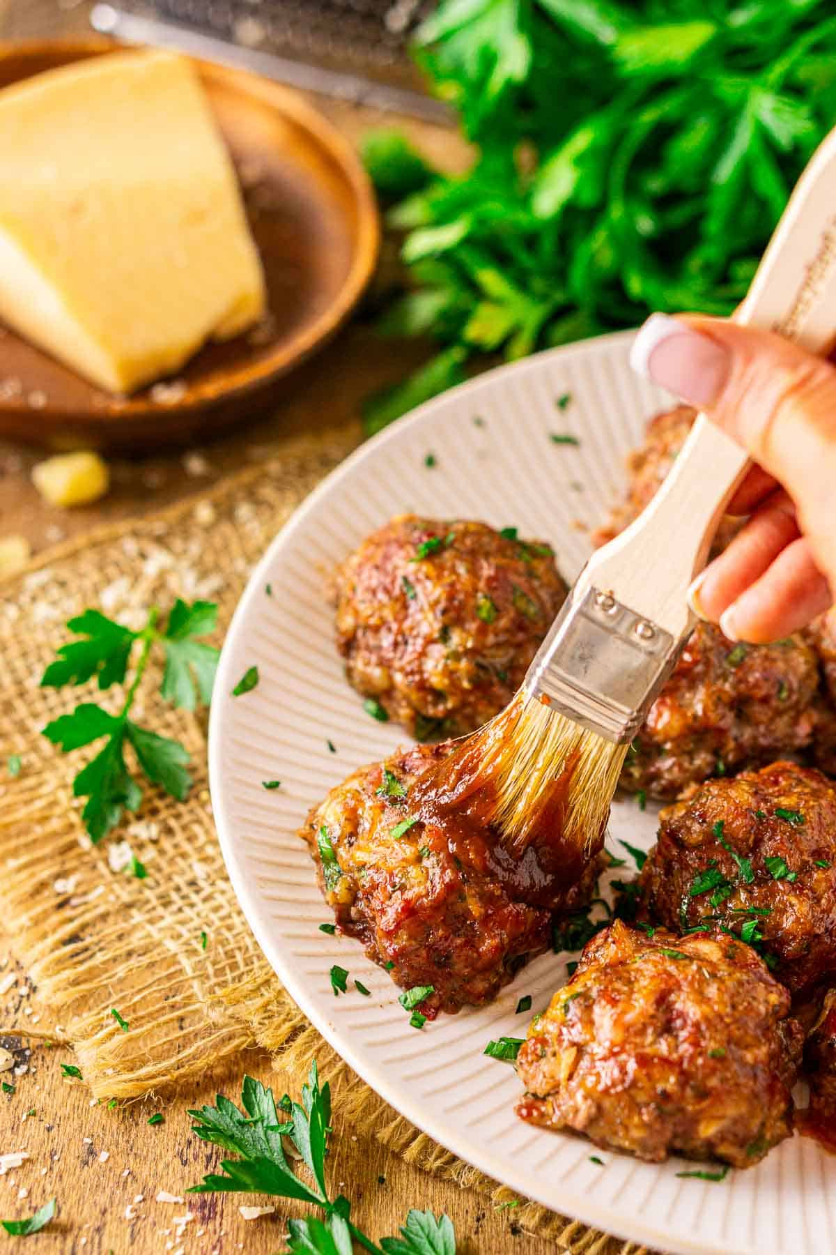 A hand brushing a smoked meatball with BBQ sauce on a white plate on top of burlap with a sprig of parsley in front.