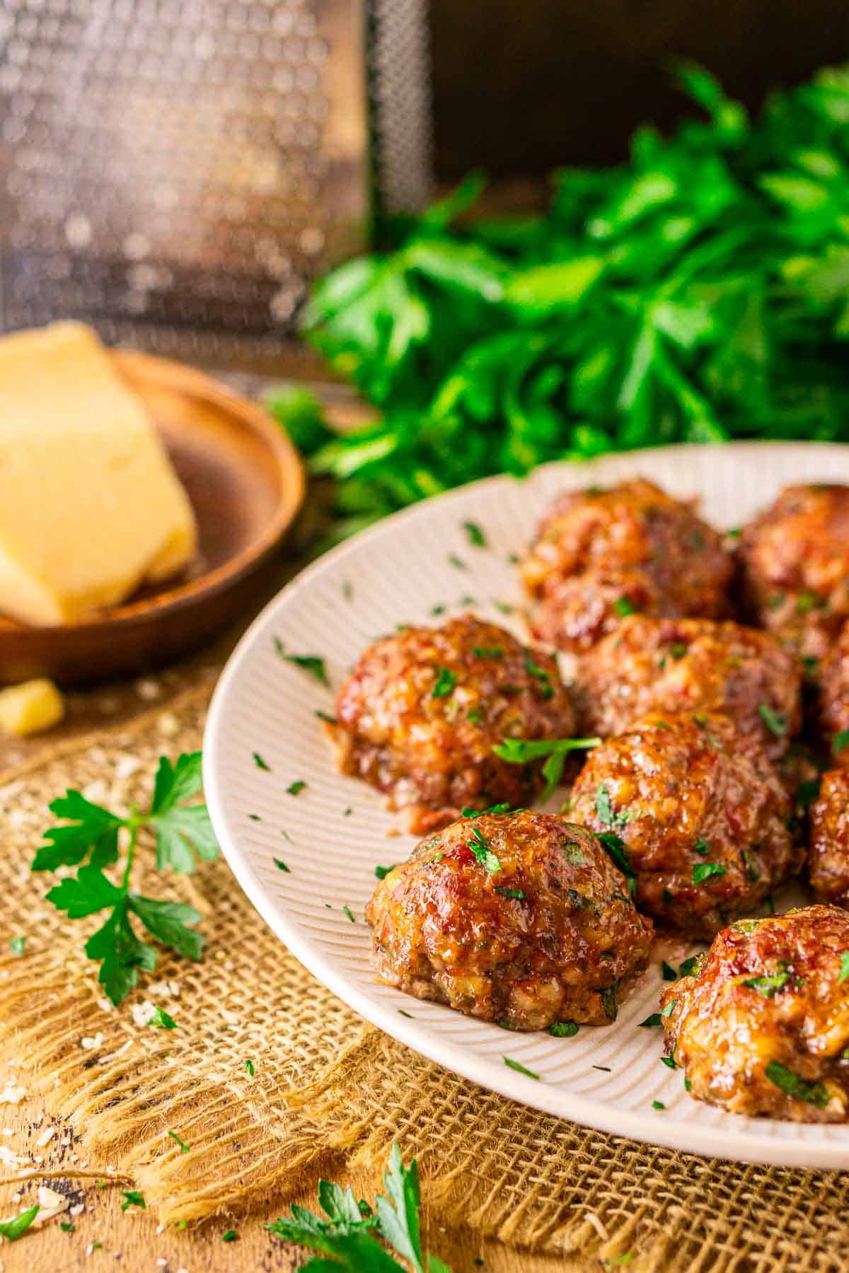 A close-up view of the smoked meatballs on a white plate with sprigs of parsley to the left and a wedge of Parmesan in the background with a cheese grater.