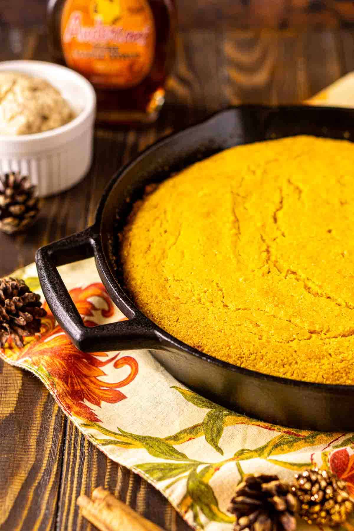 The sweet potato cornbread on a fall napkin with decor around it and a bottle of maple syrup in the background.