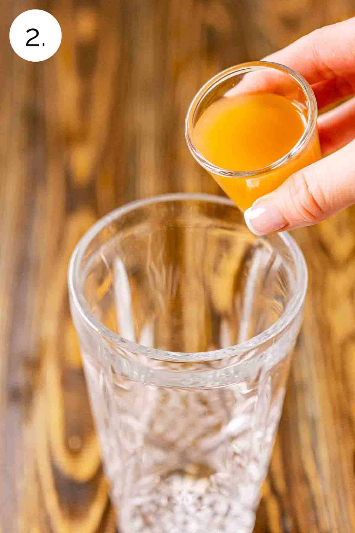 A hand pouring a shot glass filled with apple cider into a clear crystal cocktail shaker on a brown wooden surface.