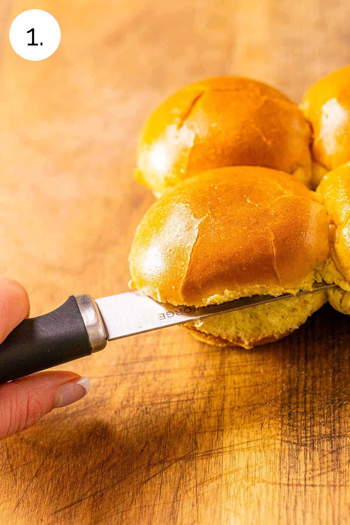 A serrated knife cutting the slider buns in half in a horizontal motion on a brown cutting board.