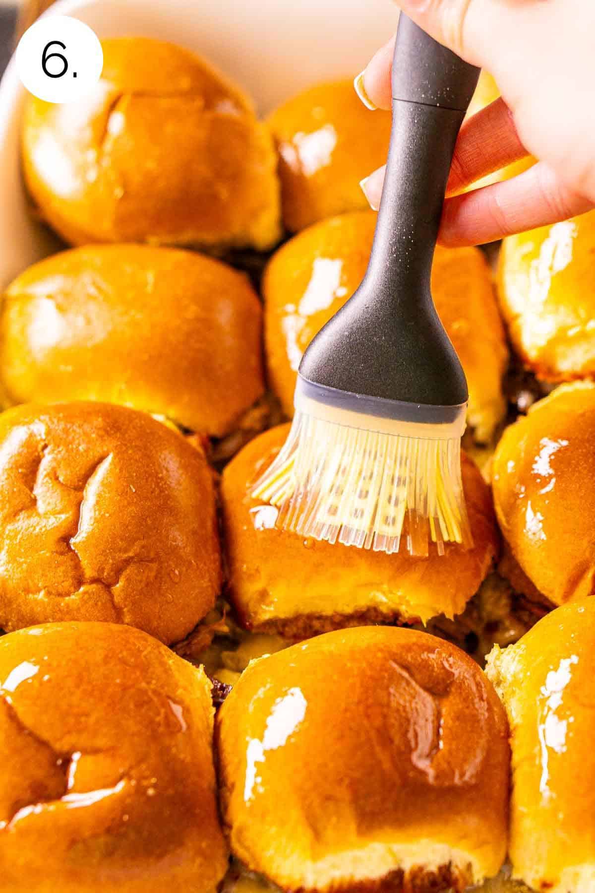 A hand coating melted butter on the slider buns with a silicone brush after they baked in the oven.