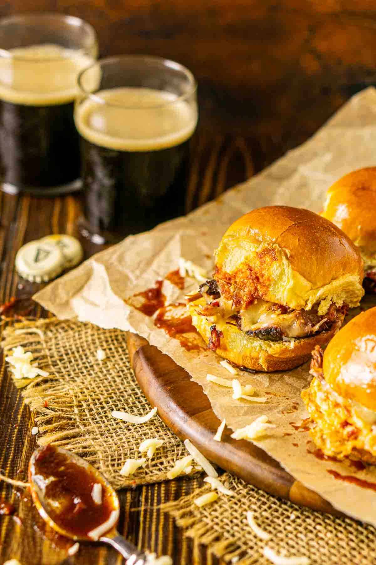 The brisket sliders on a brown wooden plate with a spoonful of BBQ sauce in front and two beers in the background.